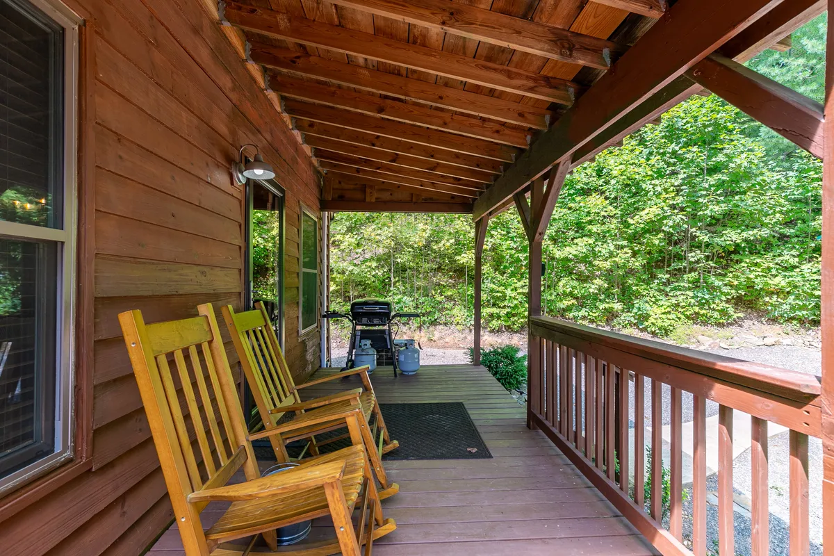 The porch features classic wooden rocking chairs, a sturdy railing, and lush green foliage. The perspective suggests a tranquil space to relax, listen to the creek, and spot wildlife, with direct access to the surrounding woods visible beyond the porch.