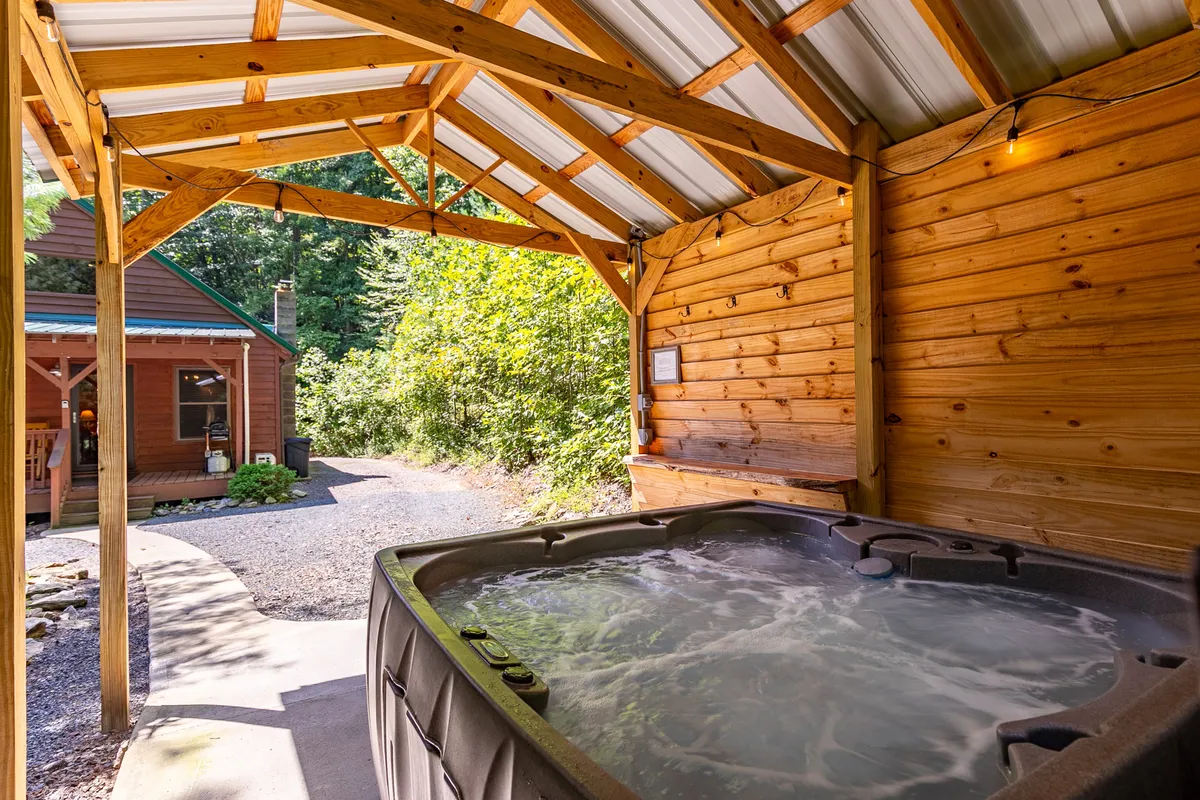 A serene view of a hot tub nestled under a wooden structure, surrounded by trees and a path leading to the cabin. The scene evokes evening soaks under a canopy of lights and stars, closely connected to nature.