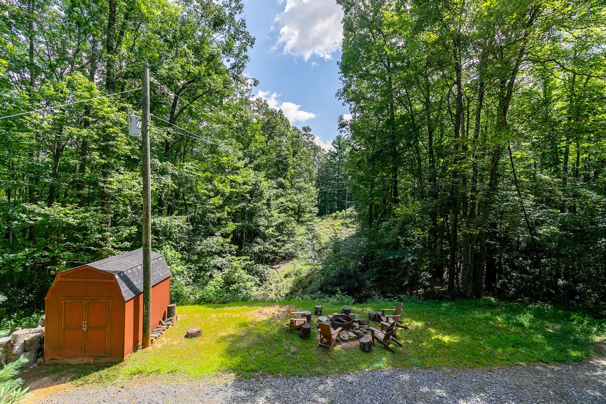 An exterior scene showing a fire pit surrounded by wooden seating, set amidst lush greenery and a charming red shed. The arrangement invites guests to gather for campfires, stories, and s’mores under the open sky.