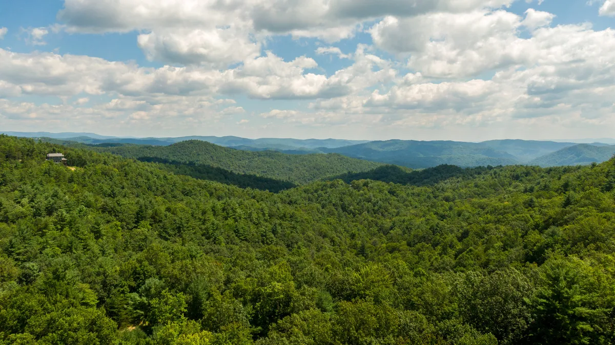 A breathtaking panoramic view featuring vast mountain ranges blanketed in dense forest beneath a cloud-dotted sky. The expansive wilderness invites exploration, while the untouched landscape underscores the sense of seclusion and privacy.