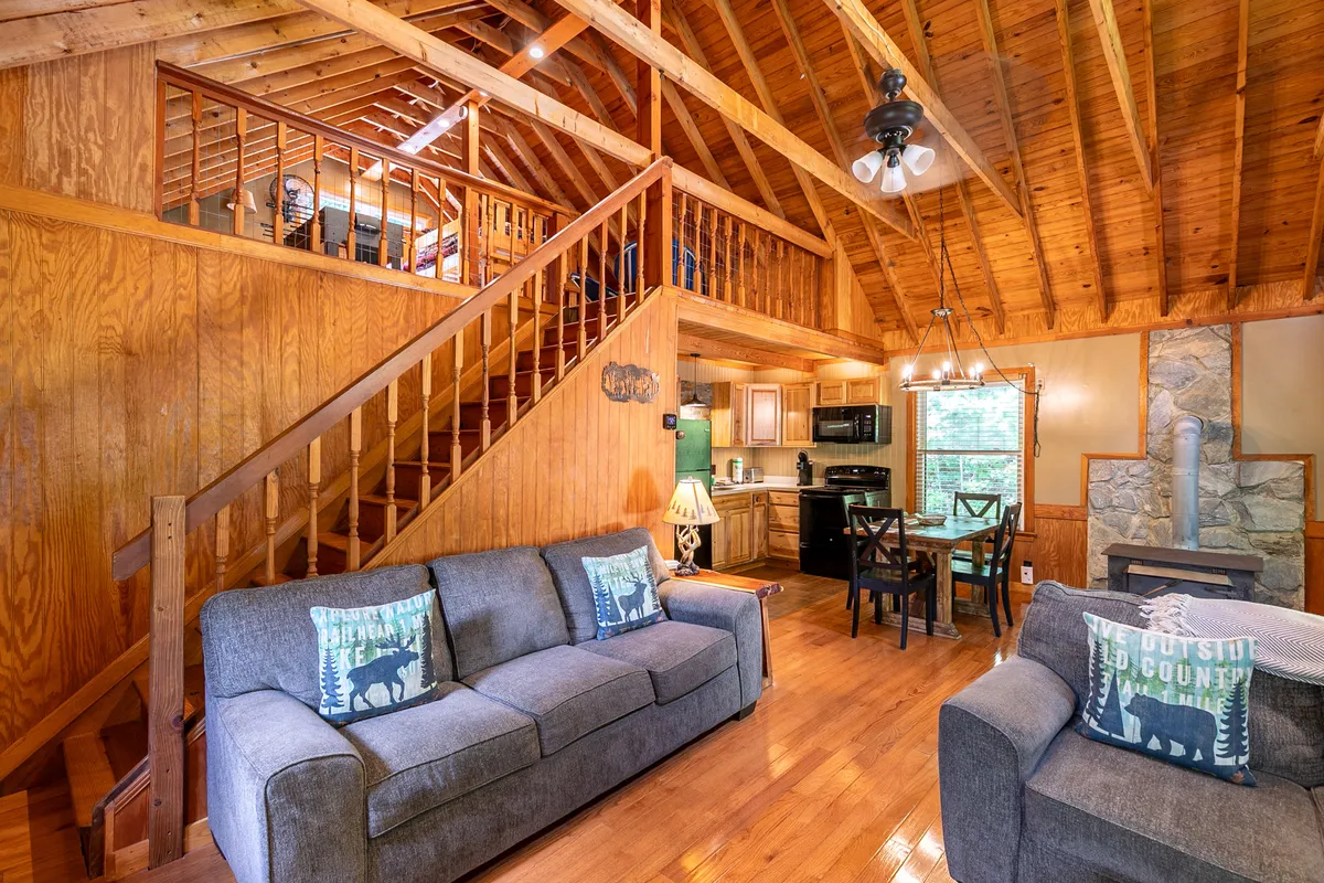 A warm, inviting interior space with a cozy sofa, decorative pillows, a staircase, dining area, and a stone fireplace. The layout hints at thoughtful amenities and comfortable spaces for relaxation and family gatherings, creating a homely atmosphere in the heart of the wilderness.