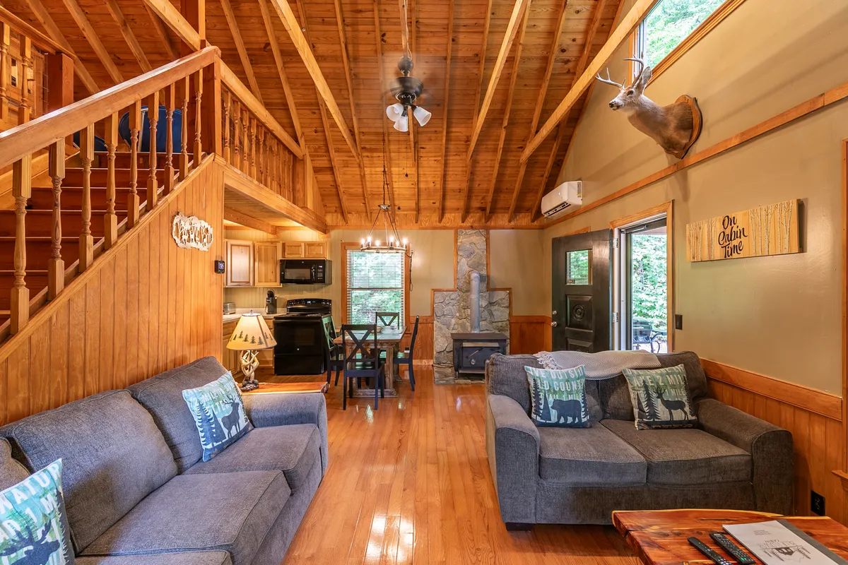 This interior image showcases a welcoming living room with comfortable sofas adorned with decorative pillows, a rustic wooden dining table, a prominent gas fireplace, exposed wooden beams, and a mounted deer head on the wall. The space is flooded with natural light, emphasizing the cabin's airy feel.