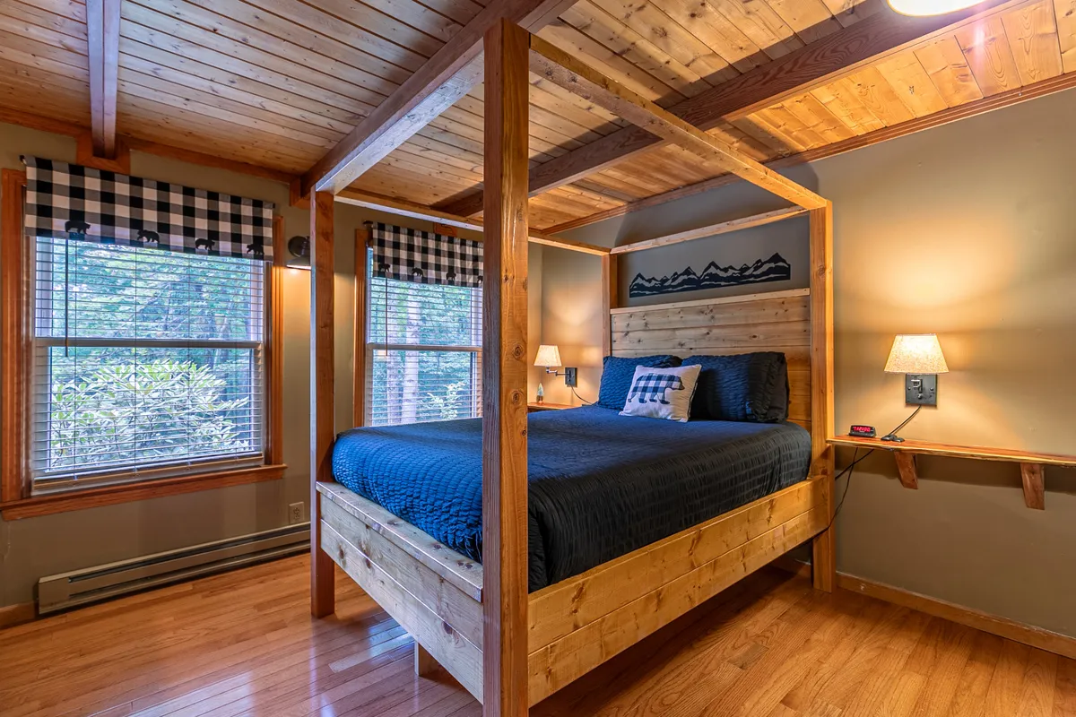 This image features a four-poster (canopy) queen bed set against a backdrop of wood-paneled walls and ceiling, with checkered curtains filtering soft natural light from the window. A nightstand with a cozy reading lamp sits beside the bed, enhancing the sense of comfort and relaxation.