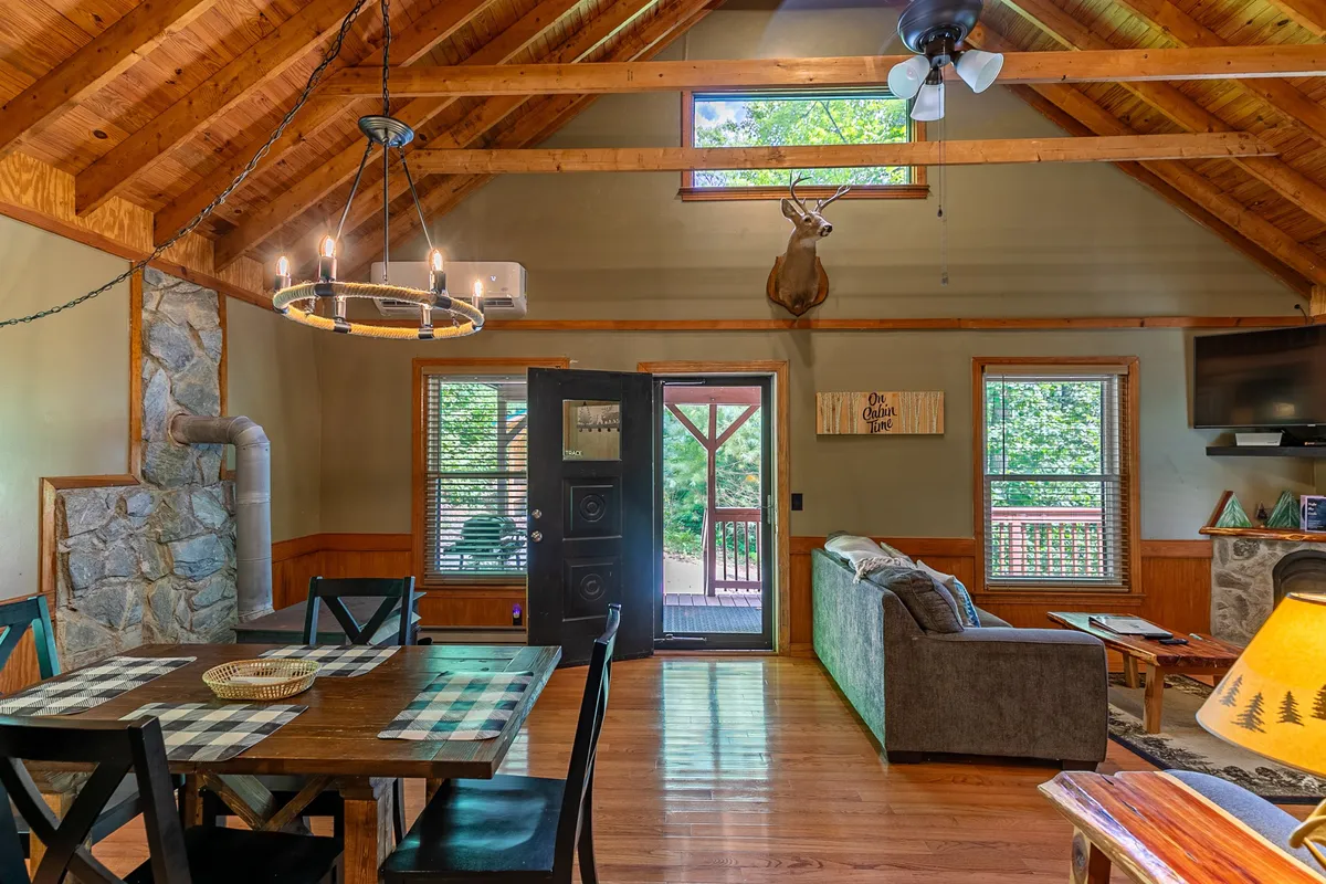 A spacious yet cozy interior with a dining table draped in a checkered cloth, a large stone fireplace, ample seating, and big windows showcasing the surrounding nature. The layout is ideal for group activities and shared meals, reinforcing the idea of family connection and versatile fun.