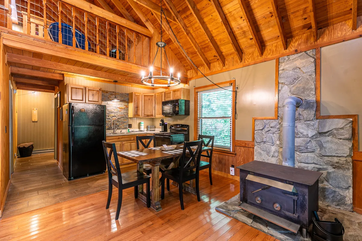 This photo features an open-concept dining and kitchen area with a central dining table, a large stone fireplace, and a fully visible, well-equipped kitchen in the background. Warm lighting and wooden finishes create a cohesive, homely feel.