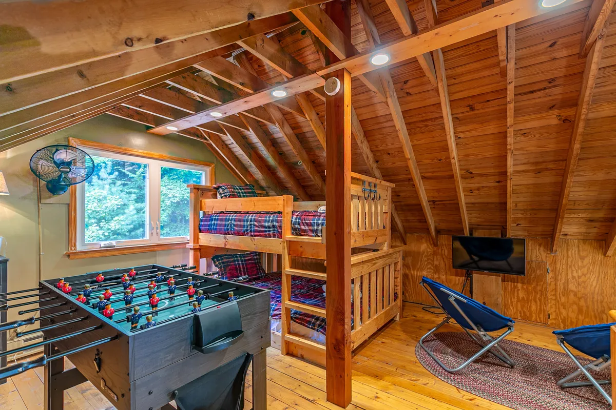 A cheerful loft space features a bunk bed (full on bottom, twin on top), a foosball table, a sunny window, and a cozy sitting area, all bathed in natural light. The atmosphere is playful and inviting, ideal for families and friends.